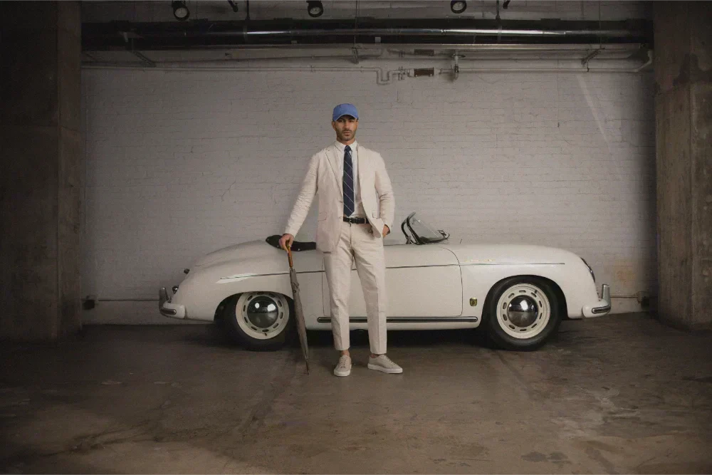 Man in a light-colored suit and Capri suede sneakers standing beside a vintage car in a garage