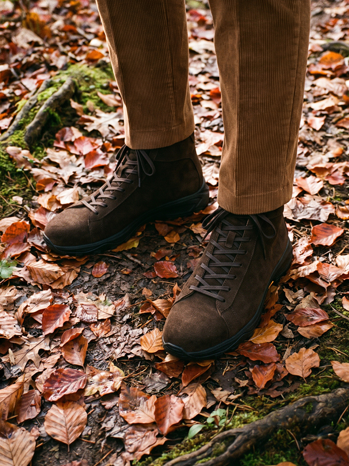 A person wearing men's brown Italian suede lace-up boots and tan corduroy pants standing on a forest floor covered in fallen autumn leaves.
