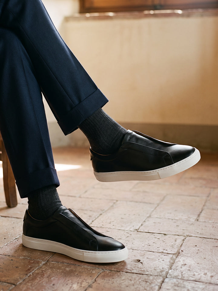 Man sitting cross legged wearing navy trousers, gray socks and men's black Italian leather slip on dress sneakers with white rubber soles.