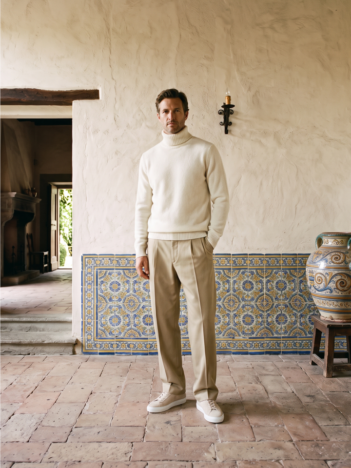 Man standing wearing khakis, a cream turtleneck and men's beige Italian suede lace-up dress sneakers with white rubber soles.