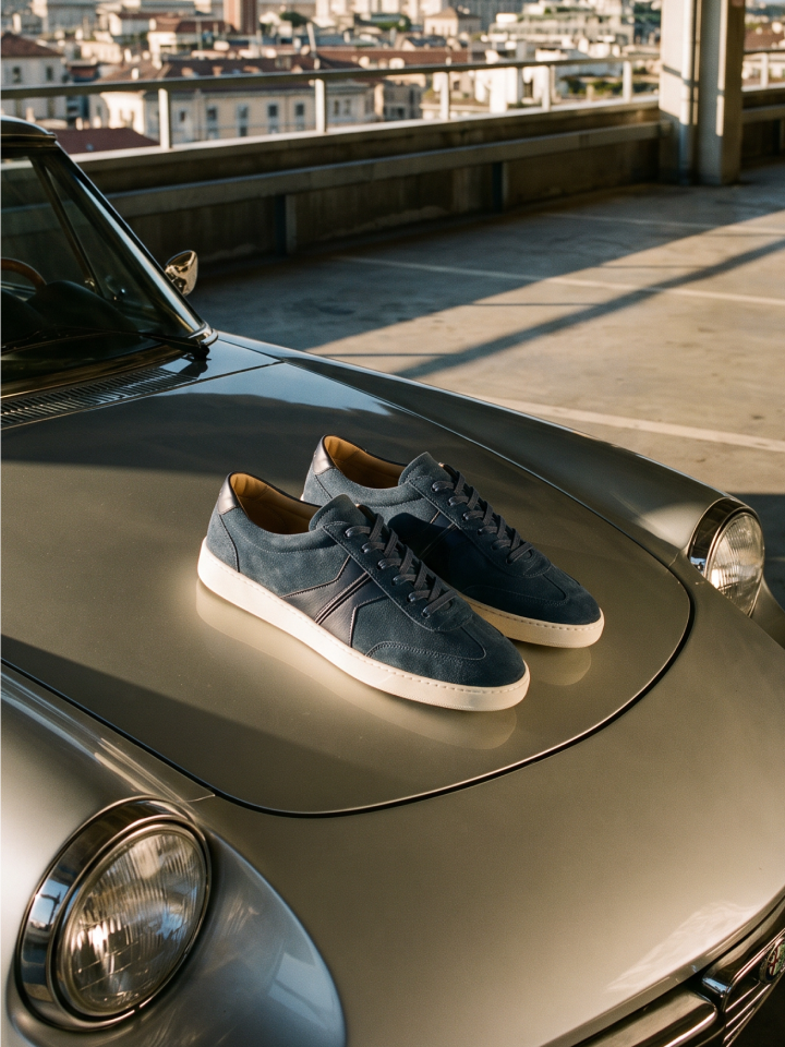 A pair of men's Italian navy blue suede lace-up sneakers with white rubber soles and tonal side stripes, styled on the hood of a vintage metallic car in a sunlit parking structure.