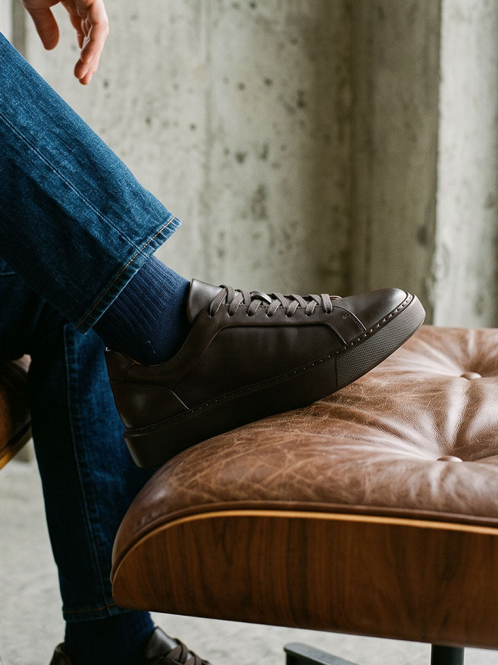 Man wearing blue jeans with navy socks while stepping on a caramel leather ottoman wearing men's brown Italian leather lace-up sneaker with tonal rubber sole.