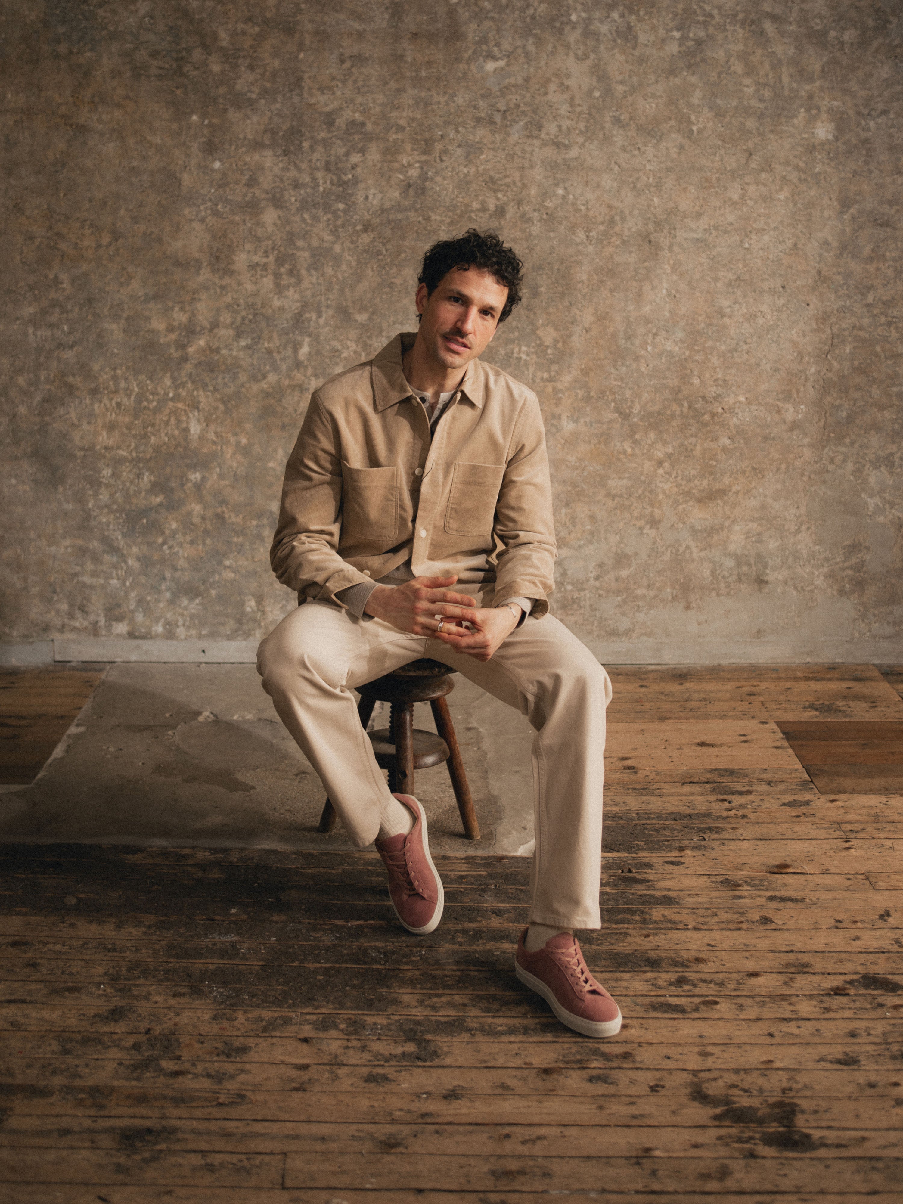 Man sitting on a stool wearing a beige button down shirt, beige chino pants, and deep blush suede sneakers with white rubber soles