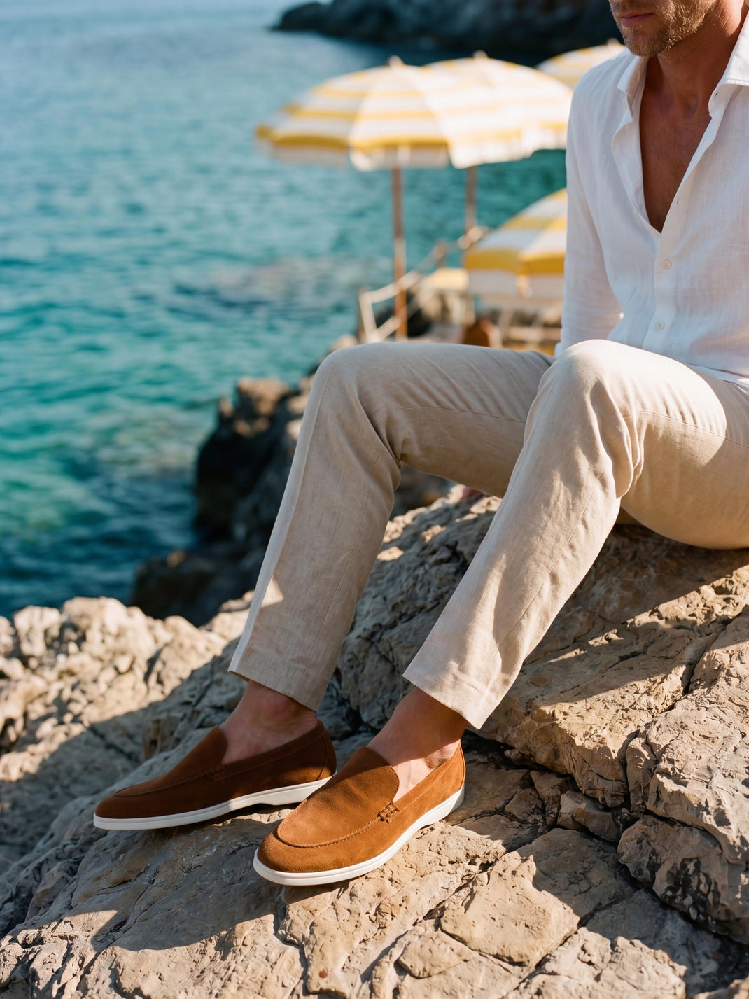 Man wearing Catania cognac suede slip-on loafers seated on rocks beside the turquoise sea