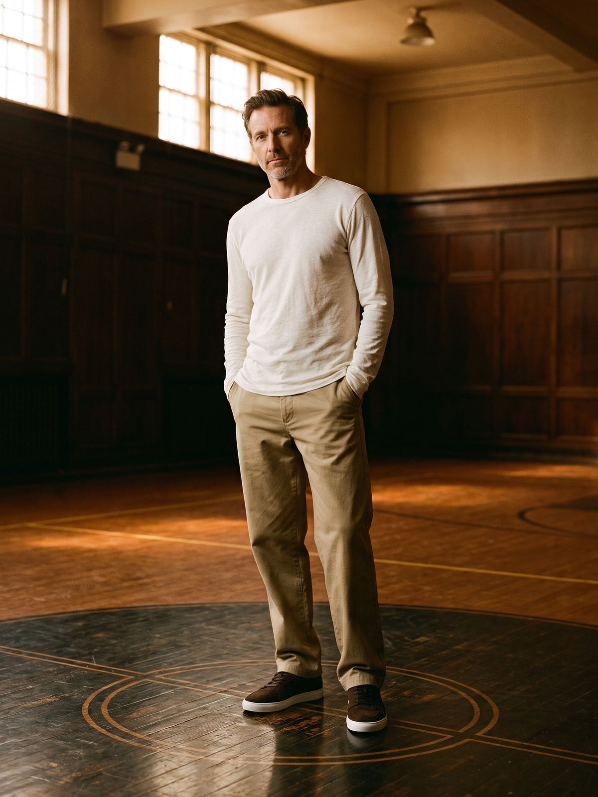 Man standing on a basketball court wearing khakis, a white long sleeve top and men