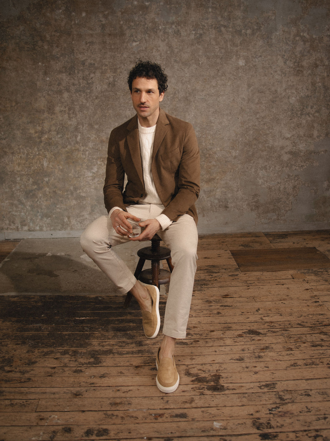 Man sitting on a stool wearing Lido in fawn, beige suede slip-on loafers in a rustic studio