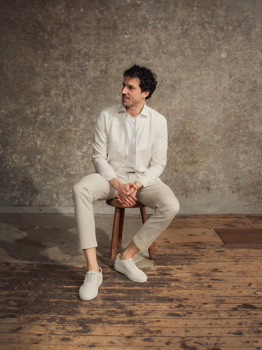 Man seated on a wooden stool wearing white suede low-top sneakers in a rustic studio