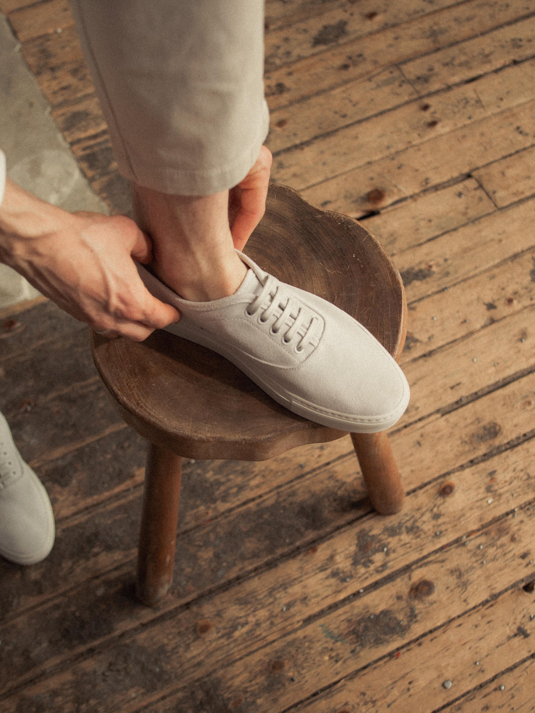 Man putting on a white suede low-top sneaker while seated on a wooden stool