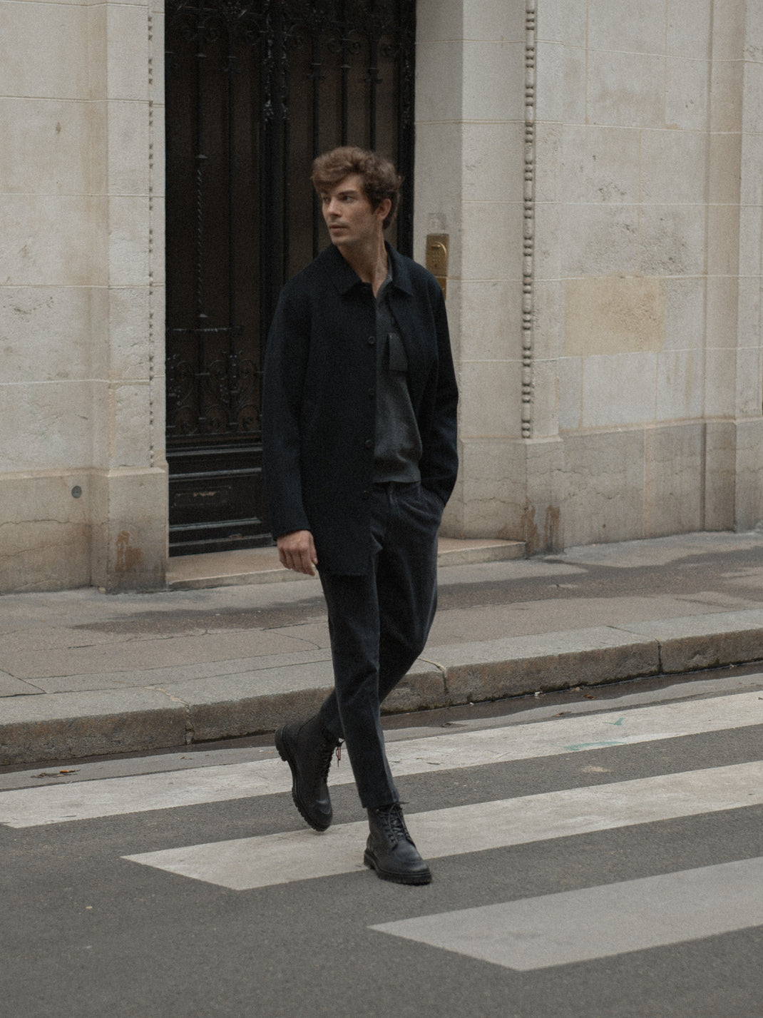 Man wearing black Milo boots walks across a city crosswalk beside a stone building