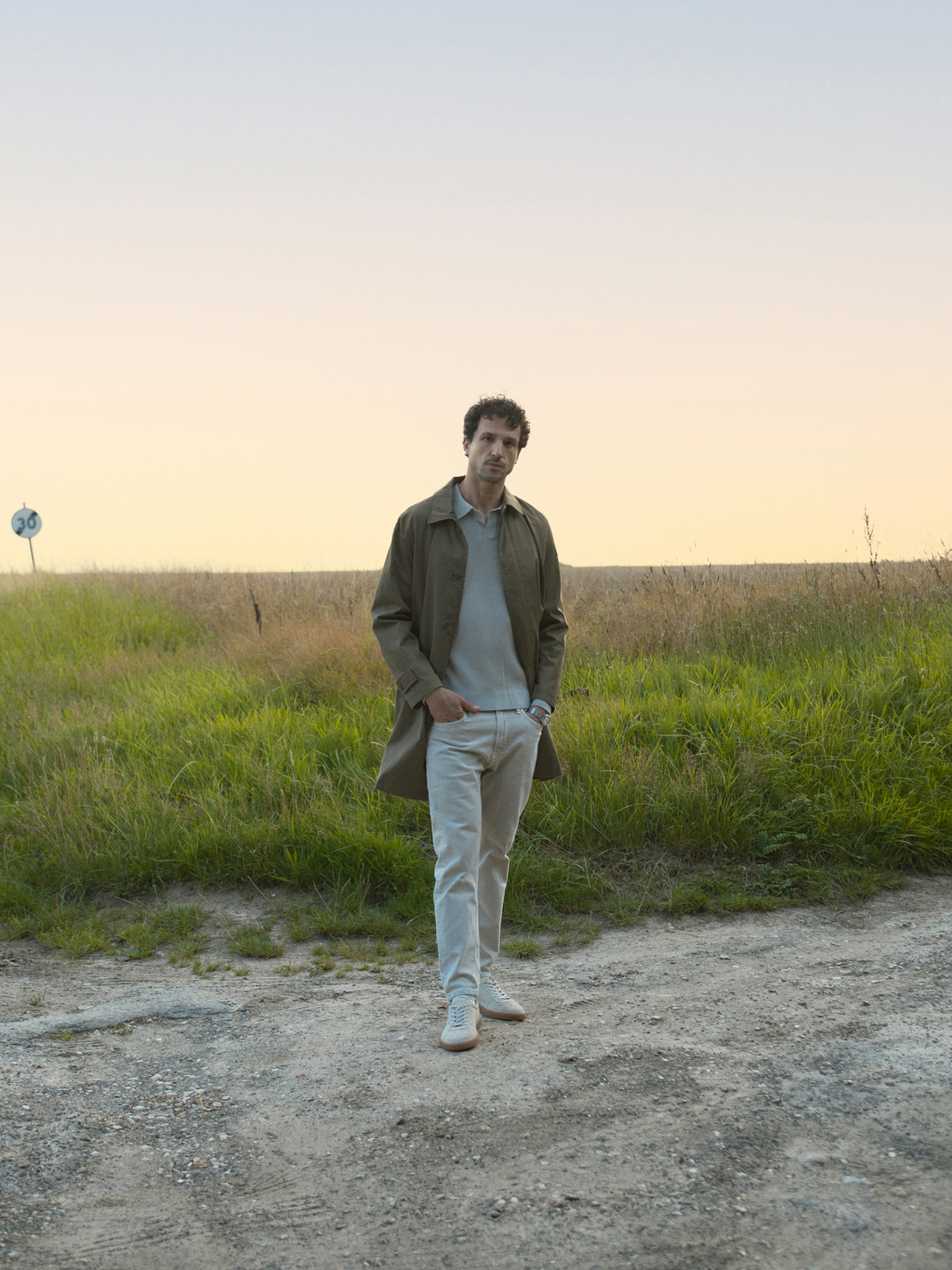 Man wearing Monza cream suede low-top sneakers standing on a country road beside a grassy field