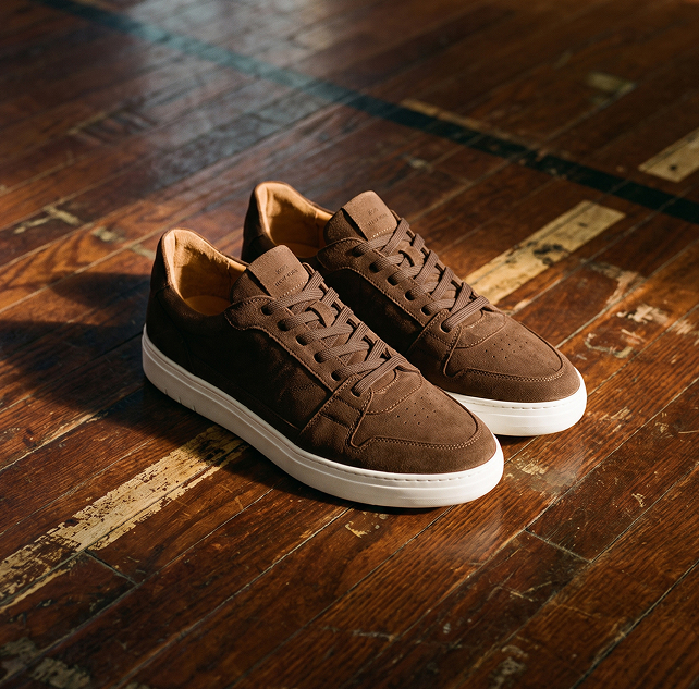 A pair of dark brown men's brown Italian suede lace-up sneakers with white rubber soles on a wooden floor.