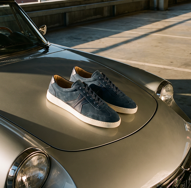 A pair of men's Italian leather dark blue suede sneakers with white rubber soles and subtle side stripes, displayed on the hood of a metallic silver car in a sunlit outdoor setting.