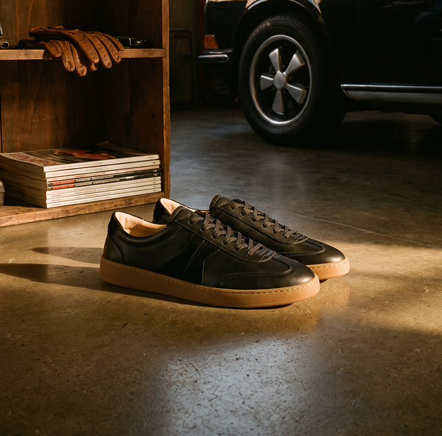 A pair of men's Italian black leather low-top lace-up sneakers with tan rubber soles, placed on a concrete floor in a garage-like setting with a wooden shelf, gloves, magazines, and part of a black car visible in the background.