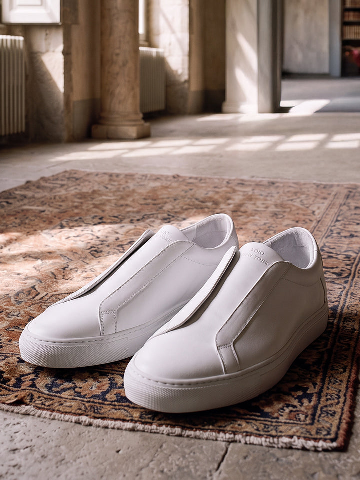 Men's white Italian leather slip on lace-less dress sneakers with white rubber soles laying on a rug in a room.