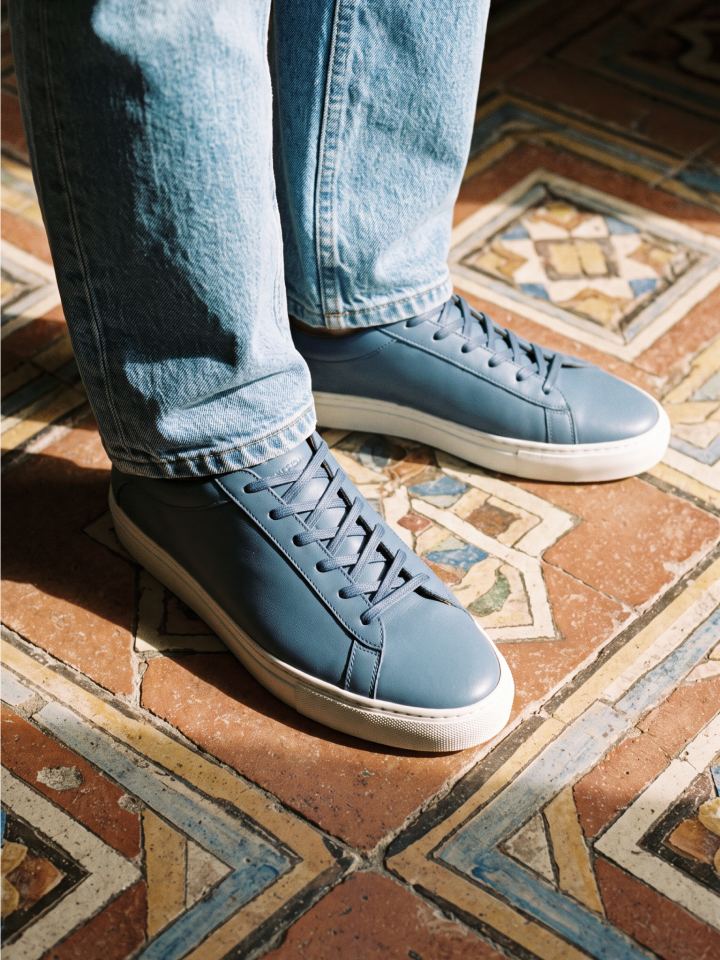 Man standing wearing men's blue Italian leather lace-up sneakers with white rubber soles and jeans.