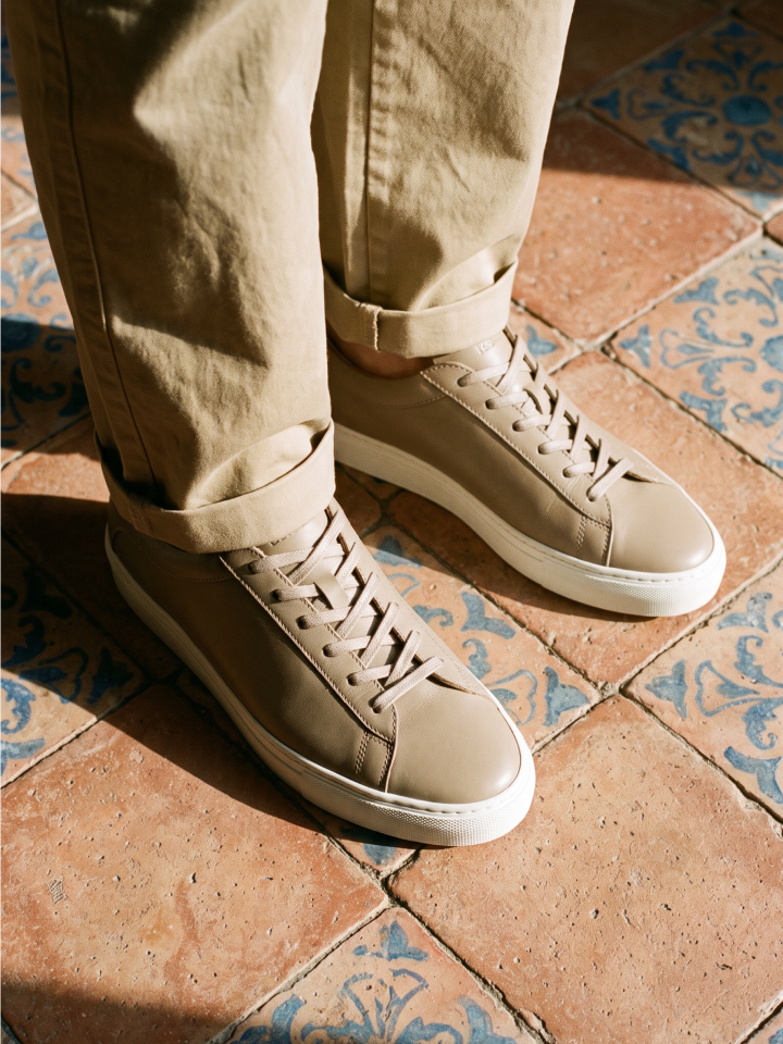 A person wearing men's beige Italian leather dress sneakers with matching beige pants, standing on terracotta tiles with blue decorative patterns.